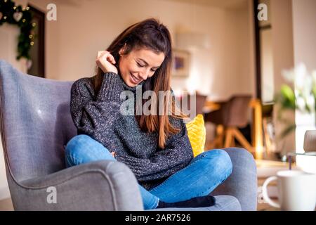 Schuss einer attraktiven lächelnden Frau mit Turtleneck-Pullover und Jeans, während sie sich zuhause im Sessel entspannen. Stockfoto