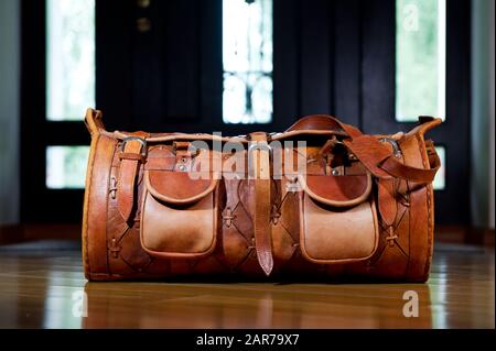 In der Nähe der Haustür eines Privathauses steht eine Ledertasche mit Sachen. Hinter der Tasche sieht man die Haustür und dahinter das Sonnenlicht Stockfoto