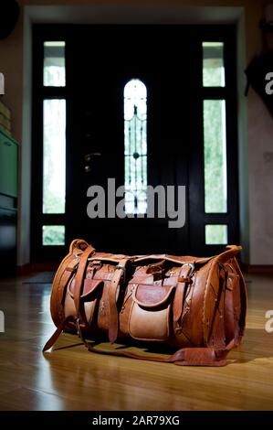 Vertikale Aufnahme einer Reisetasche aus Leder, die im Flur in der Nähe der Haustür steht, hinter der Tageslicht sichtbar ist Stockfoto