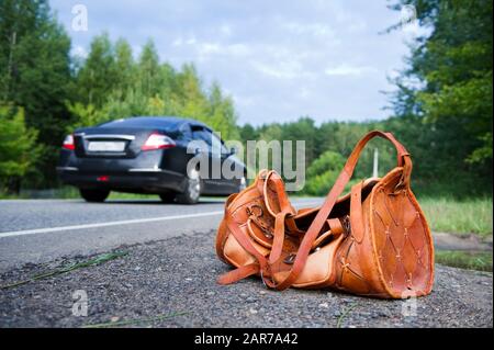Eine orangefarbene Ledertasche an einem Sommertag an der Seite einer Landstraße und ein vorbeifahrendes Auto im Hintergrund. Großer Winkel. Kopierbereich Stockfoto