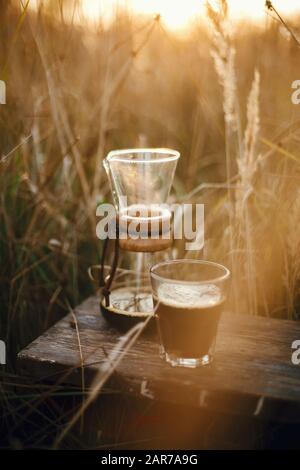 Heißer Kaffee in Glasbecher und Glaskolben im Hintergrund bei sonnigem warmem Licht in ländlichen Kräutern. Alternative Kaffeezubereitung im Freien auf Reisen. Atmosphärenr Stockfoto