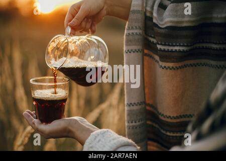 Alternative Kaffeezubereitung auf Reisen. Die Hipperin gießt frischen heißen Kaffee in einer gläsernen Tasse, im Hintergrund des sonnigen warmen Lichts in ländlichen Kräutern. Atmospheri Stockfoto