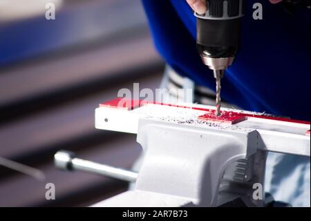 Meister in einem blauen T-Shirt und grauen Handschuhen bohrt ein in einem Schraubstock eingespanntes Metallstück, Arbeiter auf einer Werkbank, Nahaufnahme Stockfoto
