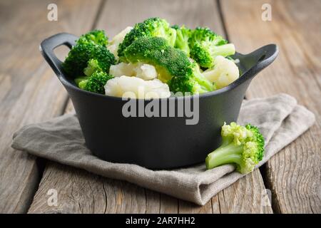 Schwarzer Topf mit gekochtem Brokkoli und Blumenkohl auf Holztisch. Stockfoto