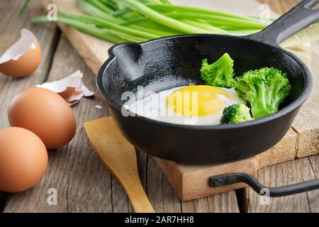 Gebratenes Ei mit Brokkoli auf einer eisernen Bratpfanne. Grüne Zwiebel auf dem Schneidbrett. Stockfoto