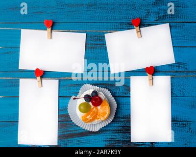 Weißes viereckiges Blatt Papier wird von roten Holzkleidern in Form von Herzen auf trendiger blauer Holzoberfläche mit köstlichem pavlova-kuchen befestigt Stockfoto