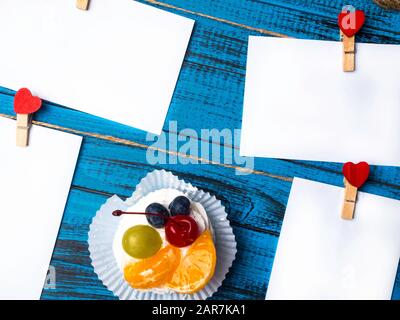 Weißes viereckiges Blatt Papier wird von roten Holzkleidern in Form von Herzen auf trendiger blauer Holzoberfläche mit köstlichem pavlova-kuchen befestigt Stockfoto