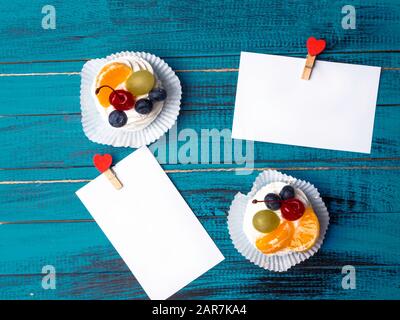 Weißes viereckiges Blatt Papier wird von roten Holzkleidern in Form von Herzen auf trendiger blauer Holzoberfläche mit köstlichem pavlova-kuchen befestigt Stockfoto