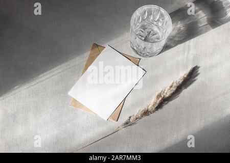 Schreibwaren Stillleben Szene. Glas Wasser, Trockenrasen und Handarbeitshülle auf grauem Tischhintergrund bei Sonnenlicht. Leere Papierkarte, Einladung Mockup sc Stockfoto