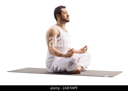 Junger Mann, der auf einer Trainingsmatte mit gekreuzten Beinen sitzt und geschlossene Augen isoliert auf weißem Hintergrund Stockfoto