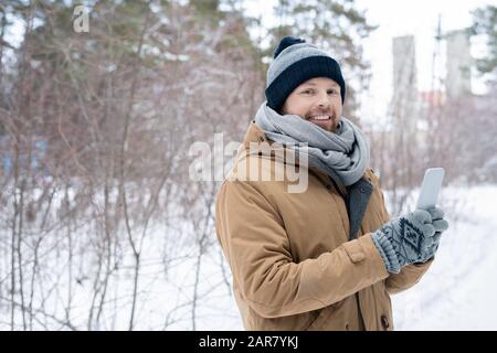 Fröhlicher junger Mann in warmer Winterkleidung, der Smartphone vor sich hält Stockfoto