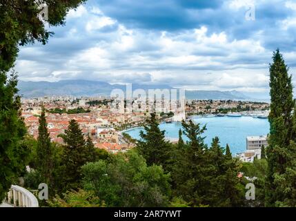 Split, Kroatien: Blick über die Altstadt mit bunten Gebäuden, Riva Promenade, Palmen und Bucht vom Hügel Marjan. Berge im Hintergrund, Bäume im Hintergrund Stockfoto