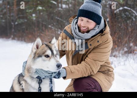 Junger lächelnder Mann im Winter, der reinrassigen sibirischen Husky-Hund kuschelt Stockfoto