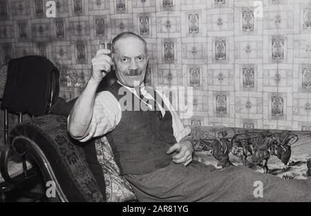 1950er Jahre, historisch, Cheers!....inside ein Zimmer, ein älterer Mann in einem Hemd und Krawatte mit einer Taille, entspannend auf einer Chaiselongue mit einem Glas seines hausgemachten Met, auch bekannt als Honigwein. Ein altes alkoholisches Getränk, das durch Gärung von Honig mit Wasser und manchmal Früchten, Gewürzen, Körnern oder Hopfen hergestellt wird, Mead soll zu einem bestimmten Zeitpunkt im Neolithikum mit der Verwendung der Gärung Lebensmittel Konservierung entdeckt worden sein. Stockfoto