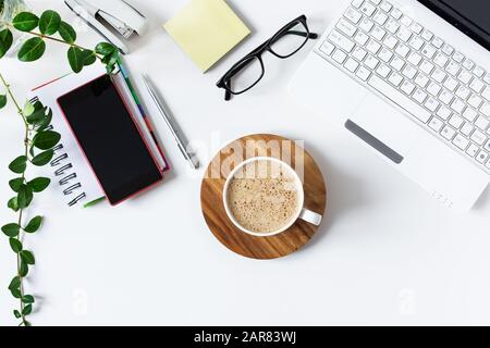 Arbeitsbereich mit Laptop, Tagebuch und einer Tasse Kaffee auf weißem Hintergrund. Eleganter Schreibtisch in weißer Farbe mit Blumentopf. Flaches Lay. Draufsicht. Stockfoto