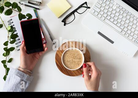 Moderner Arbeitsplatz in weißer Farbe mit Frauhand über Handy und Kaffeetrinken im Büro. Draufsicht. Flaches Lay. Stockfoto