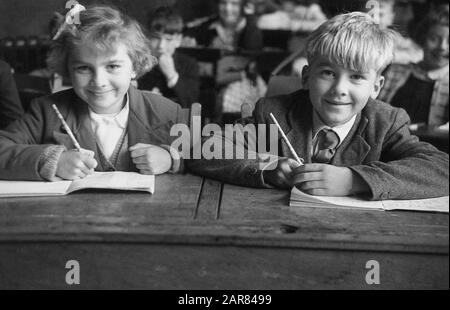 1955, historisch, sitzen zwei junge Grundschulkinder nebeneinander an einem Holzschreibtisch der Epoche in einem Klassenzimmer, die mit ihren offenen Büchern und Bleistiften in der Hand lernen möchten, England, Großbritannien. Stockfoto