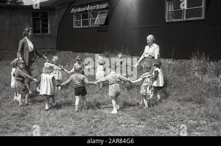 1948, kleine Flüchtlingskinder aus Osteuropa, die mit ihren Betreuerinnen außerhalb ihrer Unterbringung oder ihres Wohnzimmers ein "Ring a Ring a Roses"-Spiel spielen, vorgefertigte nissen-Hütten, England, Vereinigtes Königreich. Das polnische Neuansiedlungsgesetz von 1947 war ein Gesetz des britischen Parlaments, das mehr als 200.000 polnische Personen erlaubte, im Vereinigten Königreich zu bleiben. Stockfoto