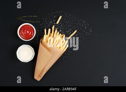 Leckere pommes Frites mit Saucen, Salz und Kräutern auf dunklem Hintergrund. Pommes frites im Papierkegel. Horizontal mit Platz für Text. Stockfoto