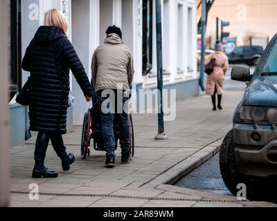 Moskau, Russland - 17. Januar 2020: Ein Mann trägt eine Person im Rollstuhl entlang des mit Fliesen gepflasterten Gehwegs. Eine Frau kümmert sich um sie. Geparkte Autos i. Stockfoto