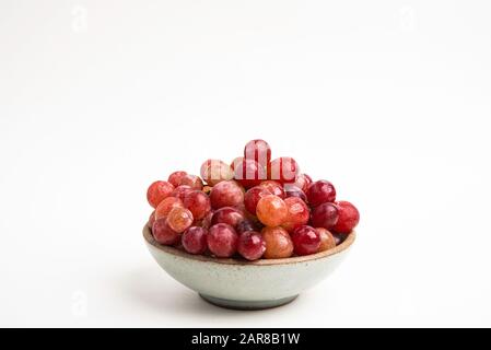 Ein Haufen frischer roter Trauben, die auf einer kleinen Keramikschale auf einem einfarbigen weißen Hintergrund gebündelt sind. Stockfoto