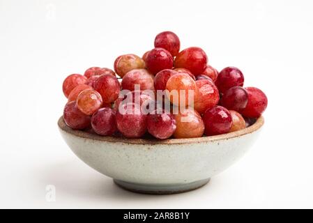 Ein Haufen frischer roter Trauben, die auf einer kleinen Keramikschale auf einem einfarbigen weißen Hintergrund gebündelt sind. Stockfoto
