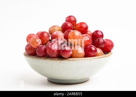 Ein Haufen frischer roter Trauben, die auf einer kleinen Keramikschale auf einem einfarbigen weißen Hintergrund gebündelt sind. Stockfoto