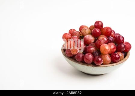 Ein Haufen frischer roter Trauben, die auf einer kleinen Keramikschale auf einem einfarbigen weißen Hintergrund gebündelt sind. Stockfoto