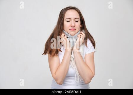 Charmantes Mädchen in einem weißen T-Shirt auf grauem Hintergrund hält an einem um seine Kehle gewickelten Schal fest. Konzeptprävention und Behandlung von Erkältungen und Grippe Stockfoto