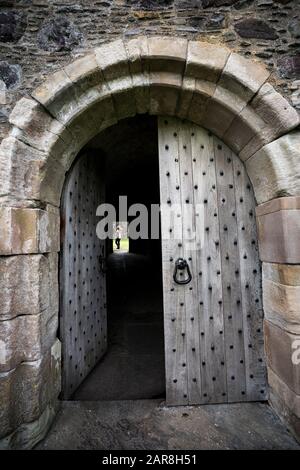 Fernblick auf eine weibliche Touristin durch die gewölbte Tür hat eine massive verschlissene Holztür, die von Steinziegeln umgeben ist, Dunstaffnage Castle aus t Stockfoto