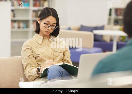 Porträt des asiatischen Teenager-Mädchens, das in der College-Bibliothek studiert und in Notizbuch schreibt, während er auf einer bequemen Couch sitzt, Kopieraum Stockfoto