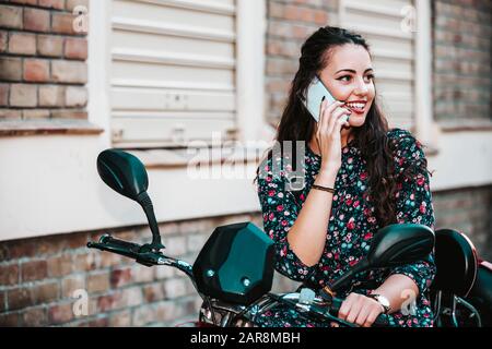 Fröhliche, fröhliche junge Frau, die auf dem Telefon auf der Straße spricht, lächelndes Mädchen, das auf ihrem Roller sitzend per Handy telefoniert. Schöne Dame Stockfoto
