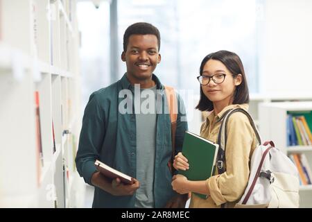Taille hoch Porträt von zwei internationalen Studenten, die Kamera und lächeln, während sie in Regalen in der College-Bibliothek posieren, Kopierraum Stockfoto