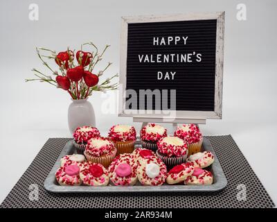 Happy Valentine's Day Letter Board mit Herzblumen-Anordnung und einem Rustikalen Tablett Voller Dekorierter Cupcakes und Thumbprint-Plätzchen, die in Frosti bedeckt sind Stockfoto