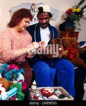 In einem Los Angeles Hospiz jubelt ein Freiwilliger ein sterbendes AIDS-Opfer an, indem er ihm hilft, Teddybären für Weihnachtsgeschenke zu dekorieren. Handwerkskit im Vordergrund notieren. Stockfoto