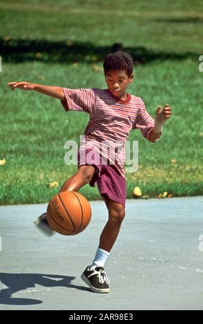 Ein afroamerikanischer zwölf Jahre alter Junge balanciert auf einem Fuß, während er während eines Outdoor-Spiels in einem Mission Viejo, CA, Park einen Basketball fängt. Stockfoto