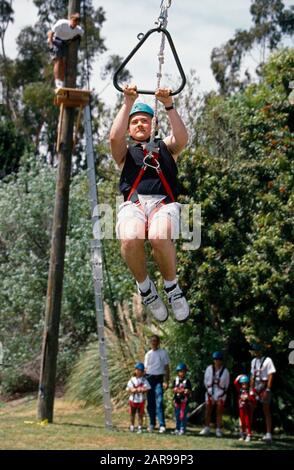 Ein Teenager, der einen harten Hut und einen Strang trägt, rutscht in einer Vertrauensbaustunde in Irvine, CA, einen Reißverschlussdraht hinunter, wie andere Schüler und Lehrer sehen. Stockfoto