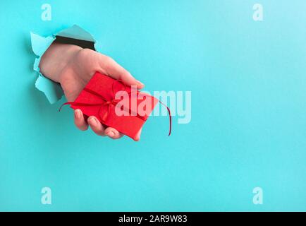 Die Hand einer Frau hält ein Geschenk durch ein Loch auf türkisfarbenem Hintergrund. Urlaubskonzept mit Platz für Text Stockfoto