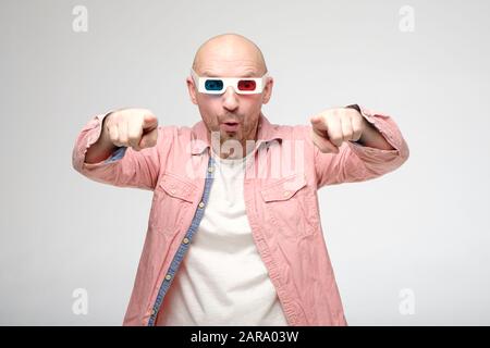 Aufgeregt, überraschter Kahlmann sieht einen Film in 3D-Brille, er stand emotional auf und zeigt einen Finger auf den Bildschirm. Stockfoto