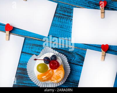 Weißes viereckiges Blatt Papier wird von roten Holzkleidern in Form von Herzen auf trendiger blauer Holzoberfläche mit köstlichem pavlova-kuchen befestigt Stockfoto