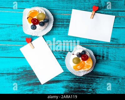 Weißes viereckiges Blatt Papier wird von roten Holzkleidern in Form von Herzen auf trendiger blauer Holzoberfläche mit köstlichem pavlova-kuchen befestigt Stockfoto