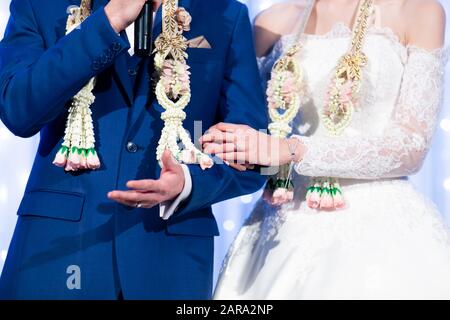 Brautpaar in formellen Kostümen mit Blumengirlanden, nervös aussehend, wurden auf der Bühne in der traditionellen thailändischen Hochzeitsfeier interviewt Stockfoto