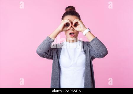 Porträt des staunenden brünetten Teenagermädchens mit bunter Frisur in lässiger Kleidung, das in Ferngläsern durch die Finger blickt, überrascht von dem, was Stockfoto