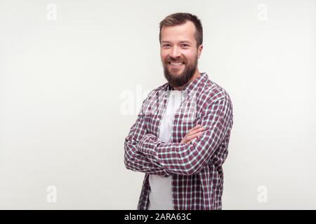 Portrait des fröhlichen gutmütigen bärtigen jungen Mannes in legeren Krawallen, die Arme kreuzen und mit freundlichen Grüßen an der Kamera lächeln, zufrieden aussehend w Stockfoto