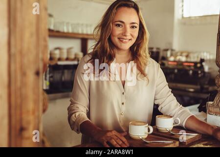 Porträt einer jungen Frau, die hinter der Theke eines Kaffeehaus arbeitet. Lächelnde Barista der Frau, die Kaffee serviert. Stockfoto