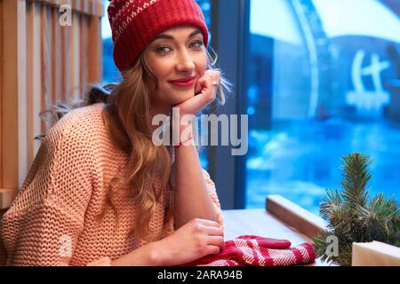 Schöne kaukasische blonde Frau in roter Strickmütze hinter Cafeteria-Fenster, die an einem wunderbaren Weihnachtstag eine warme Latte trinkt. geschweifte europäer Stockfoto