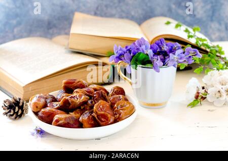 Schüssel mit süßlich leckeren getrockneten Datumsfrüchten in einer Komposition mit violetten Blumen und Vintage-Büchern. Ramadan, traditionelle Küche. Stockfoto