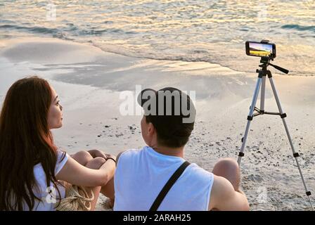 Boracay Island, Provinz Aklan, Philippinen: Kleines Paar aus Filipino, das am Strand sitzt und den Sonnenuntergang mit dem Handy fotografiert Stockfoto