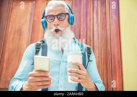 Schockierter leitender Mann, der Smartphone verwendet, während er zu Hause Musik hört und morgens Kaffee trinkt - Technologie, trendiger Lebensstil und Stockfoto