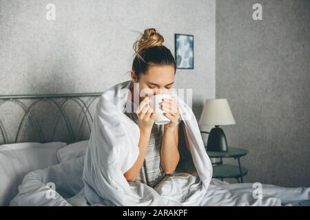 Das Mädchen trinkt ein Getränk aus einem Becher im Bett, das in eine Decke eingewickelt ist. Sie genießt, oder sie ist nicht wach oder krank. Stockfoto